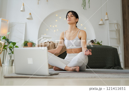 Yoga mindfulness meditation online. Woman practicing yoga with online lessons in laptop at home. Woman sitting in lotus pose meditating relaxing indoor. Girl doing breathing practice. Yoga at home Yoga mindfulness meditation online. Woman practicing yoga with online lessons in laptop at home. Woman sitting in lotus pose meditating relaxing indoor. Girl doing breathing practice. Yoga at home 121411651