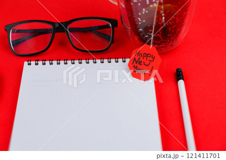 A white notebook with a black spring at the top lies in the center of the photo, next to it is a white pencil with a black eraser, transparent black-framed glasses and a mug of tea are on top, a red 121411701