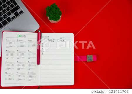 A pink diary notebook that says: Goals for 2025, inside it is a calendar for 2025 and there is a pink pen on a red background, an open laptop lies on top, part of the keyboard is visible and there is A pink diary notebook that says: Goals for 2025, inside it is a calendar for 2025 and there is a pink pen on a red background, an open laptop lies on top, part of the keyboard is visible and there is 121411702