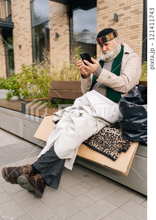 Vertical portrait of older man sitting on city bench, using typing smartphone amidst urban solitude, shedding light on poverty and isolation in aging populations. Concept of poor unhappy retirement. Vertical portrait of older man sitting on city bench, using typing smartphone amidst urban solitude, shedding light on poverty and isolation in aging populations. Concept of poor unhappy retirement. 121411743