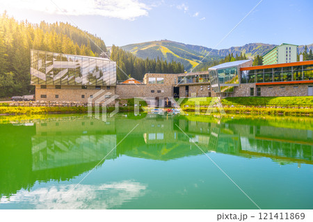 Visitors enjoy the tranquil summer atmosphere at Jasna Ski Resort in Slovakia. The lush green landscape reflects beautifully on the calm waters, highlighting the resort's modern architecture. 121411869
