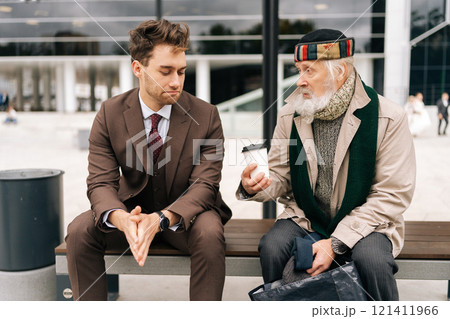 Handsome kindness businessman in formal attire attentively engages in conversation with senior homeless man holding disposable coffee cup, while seated on bench in public area, on cloudy day. 121411966