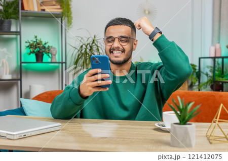 Indian Arabian man at home table use smartphone celebrating success victory winning play online game Indian Arabian man at home table use smartphone celebrating success victory winning play online game 121412075