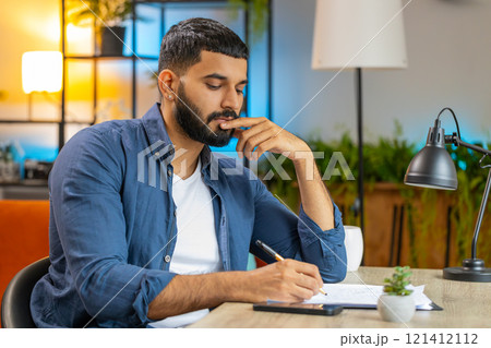 Indian man freelancer working with documents from home office analyzing reading business report Indian man freelancer working with documents from home office analyzing reading business report 121412112