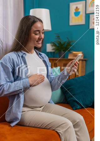 Smiling pregnant woman using smartphone making online purchases for baby sitting on home couch Smiling pregnant woman using smartphone making online purchases for baby sitting on home couch 121412159