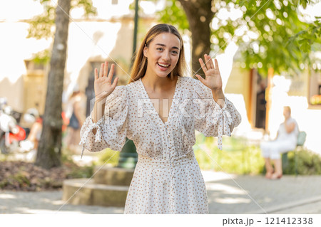 Woman in dress smiling friendly at camera waving hands gesturing hi greeting goodbye on city street Woman in dress smiling friendly at camera waving hands gesturing hi greeting goodbye on city street 121412338