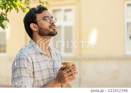 Happy Indian young man enjoying morning coffee hot drink and smiling standing on city street Happy Indian young man enjoying morning coffee hot drink and smiling standing on city street 121412346