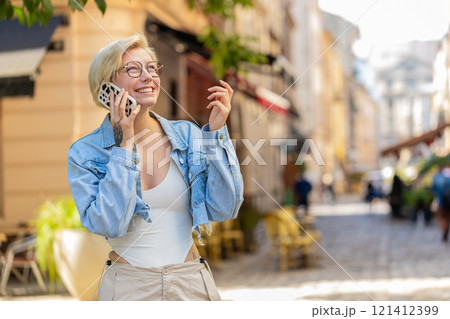 Happy young Caucasian blonde woman with short hair talking gossip on smartphone on city street 121412399