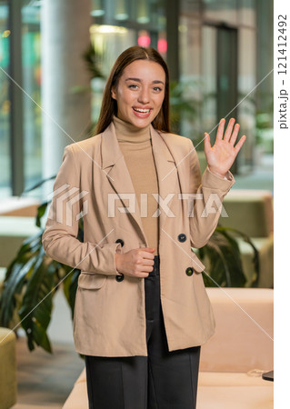 Happy young Caucasian businesswoman looking at camera and waves hand hello in office hotel lobby Happy young Caucasian businesswoman looking at camera and waves hand hello in office hotel lobby 121412492