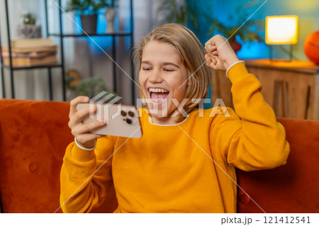 Teenager boy celebrating victory after winning mobile game on smartphone joyful jump on sofa at home 121412541
