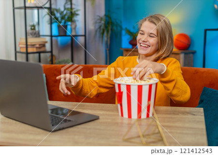 Teenager boy child watching funny video on laptop, laughing while eating popcorn on couch at home 121412543