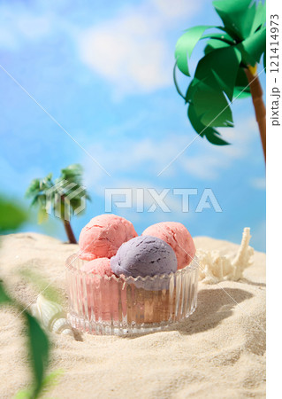 A glass bowl filled with ice cream balls is shown in a miniature summer beach for food advertising photos, surrounded by palm trees, seashells, and a coral on a sandy side. Close-up view. 121414973