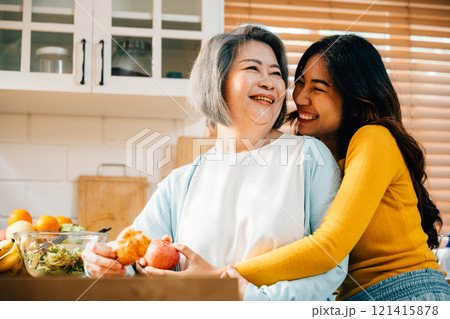 An apron-clad young Asian woman and her old mother bond over food and drinks in the kitchen. Their cheerful expressions reveal the joy of family togetherness and fun. An apron-clad young Asian woman and her old mother bond over food and drinks in the kitchen. Their cheerful expressions reveal the joy of family togetherness and fun. 121415878
