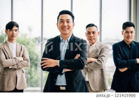 A smartly dressed businessman smiles with confidence, arms crossed in a creative office. Diverse Asian male and female colleagues stand together at a startup, symbolizing teamwork and success. 121415900