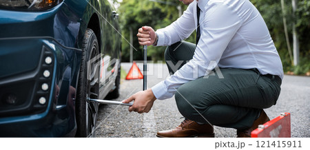 Asian young businessman unscrewing lug nuts on the wheel with a lug wrench. Handsome man wearing white shirt road assistance changing car wheel on the highway. Car mechanic changing car tyre concept. 121415911