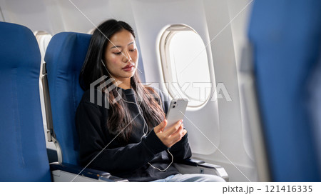 A relaxed, happy Asian woman using her smartphone and listening to music during the flight. 121416335