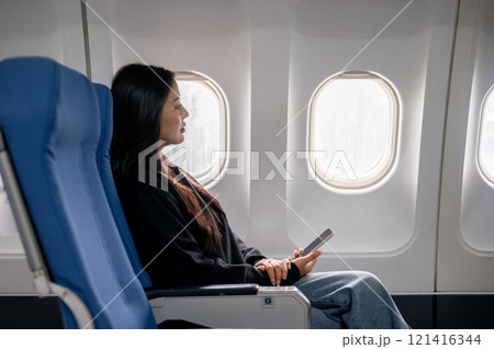 Relaxed Asian female passenger sits at a window seat, looking at the view outside during the flight 121416344