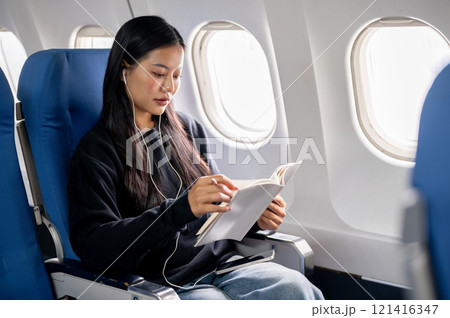 A relaxed Asian female passenger reads a book and listens to music during the flight. 121416347
