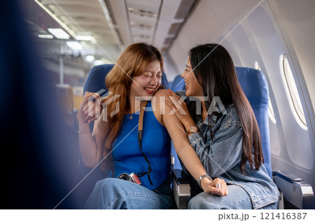 Two joyful Asian female friends, excited for their vacation, sit on a plane with happy expressions. 121416387
