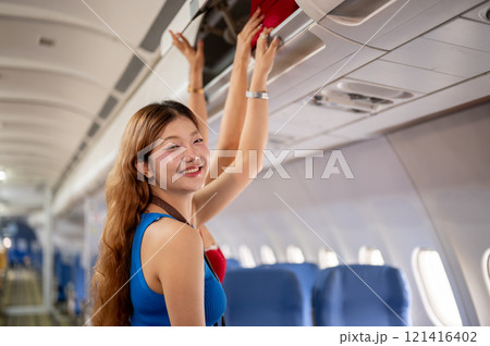 Two Asian girls place their bags in the overhead locker on the plane, with one smiling at the camera 121416402
