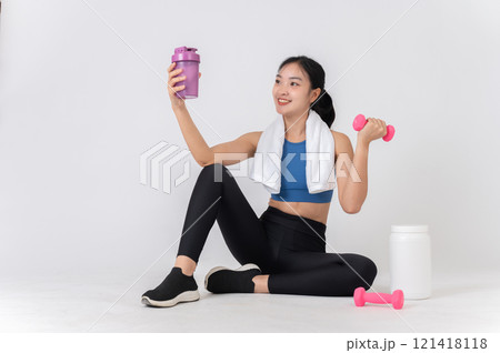 A woman in sportswear sits on a studio background, holding a dumbbell and a protein shake bottle. 121418118