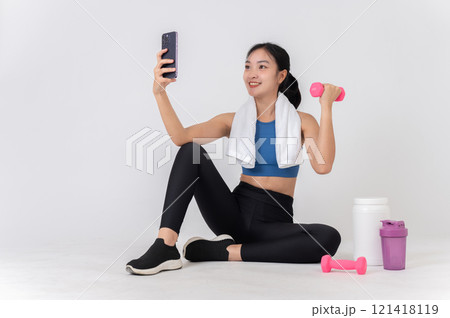 Healthy Asian woman in sportswear sits on a white studio background, taking a selfie with her phone. 121418119