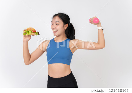 A confident Asian woman in sportswear holding a burger in one hand and a dumbbell in the other. A confident Asian woman in sportswear holding a burger in one hand and a dumbbell in the other. 121418165