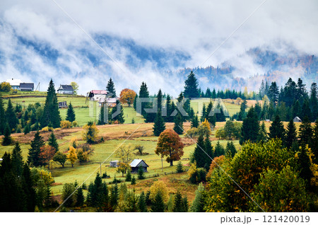 Picturesque autumn landscape in mountains. Hills dotted with colorful trees and rural houses, surrounded by evergreen forests. Mist rises from valleys, creating serene and enchanting atmosphere. Picturesque autumn landscape in mountains. Hills dotted with colorful trees and rural houses, surrounded by evergreen forests. Mist rises from valleys, creating serene and enchanting atmosphere. 121420019