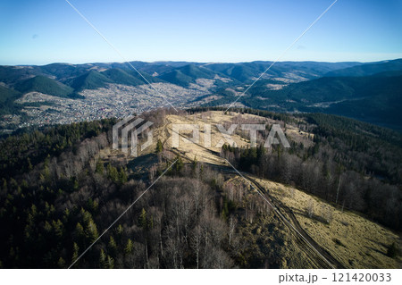 Aerial view of mountain landscape with rolling hills and dense forests. Bare trees and winding dirt trails, while background reveals village nestled in valley. Scene bathed in clear, crisp daylight. 121420033