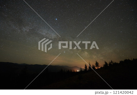 Breathtaking night sky displays dense field of stars and Milky Way over tranquil mountain landscape. Silhouetted trees and distant mountain peaks faintly illuminated by glow from below horizon. 121420042