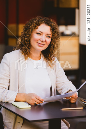 A businesswoman with curly hair is seated at an outdoor cafe, intently reading a document. 121420283