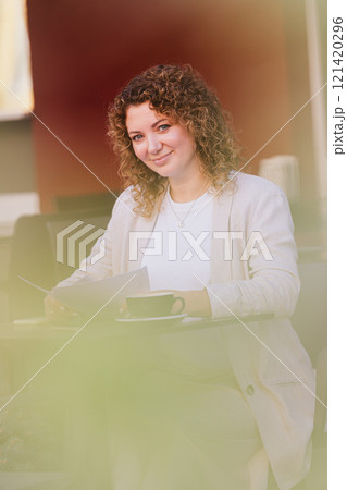 A businesswoman is seated at an outdoor cafe, enjoying a cup of coffee while reading a document 121420296