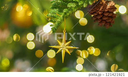 A golden star ornament hangs gracefully from a green Christmas tree branch, accompanied by a natural pinecone. Soft golden bokeh lights enhance the festive and cozy atmosphere 121421921