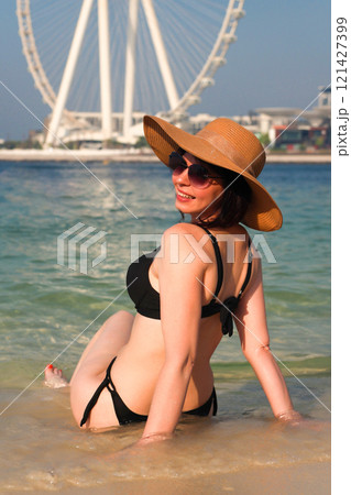 Sexy woman in hat and black bikini sitting on sand in sea waves overlooking city and amusement park Sexy woman in hat and black bikini sitting on sand in sea waves overlooking city and amusement park 121427399