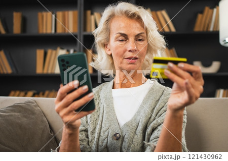 Happy middle aged woman shopping online with smartphone paying with credit card. Mature lady buying on Internet enter credit card details at home indoors. Online shopping delivery service ecommerce Happy middle aged woman shopping online with smartphone paying with credit card. Mature lady buying on Internet enter credit card details at home indoors. Online shopping delivery service ecommerce 121430962