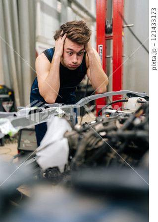 Vertical portrait of mechanic having stressful day at car repair shop, holding head in hands while looking at car engine. Confused repairman unsure of how to fix complex engine problem in garage. Vertical portrait of mechanic having stressful day at car repair shop, holding head in hands while looking at car engine. Confused repairman unsure of how to fix complex engine problem in garage. 121433173