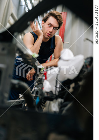 Vertical portrait of worried vehicle manager surrounded by tools and car parts. Puzzled mechanic male leaning on car in auto repair shop, looking stressed and thinking about how to fix engine. 121433177
