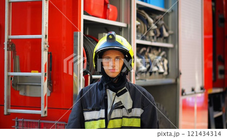 Portrait of confident female firefighter against the background of big red truck. Young firewoman in protective uniform looking at camera near a fire engine. Saving lives and heroic profession concept Portrait of confident female firefighter against the background of big red truck. Young firewoman in protective uniform looking at camera near a fire engine. Saving lives and heroic profession concept 121434314
