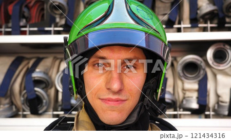 Young smiling fireman looking at camera in full equipment after fire against the background of big red truck. Portrait of happy male fireguard standing near a fire engine. Concept of heroic profession Young smiling fireman looking at camera in full equipment after fire against the background of big red truck. Portrait of happy male fireguard standing near a fire engine. Concept of heroic profession 121434316