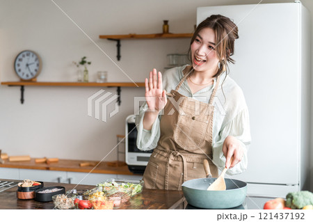 キッチンでフライパンを使うエプロン姿の女性(笑顔・手を振る) キッチンでフライパンを使うエプロン姿の女性(笑顔・手を振る) 121437192