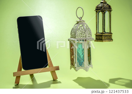 Wooden easel and mobile phone with Arabic lanterns hanging on a colored background Wooden easel and mobile phone with Arabic lanterns hanging on a colored background 121437533