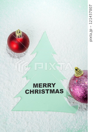 Closeup view of a text of Merry Christmas on a wooden Christmas tree ornament and Christmas balls with snow on a colored background 121437807