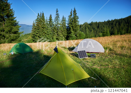 Tourist tent with solar panel and portable power station nearby in grassy field. Scene surrounded by rolling hills, tall pine trees under clear blue sky, showcasing eco-friendly camping setup. Tourist tent with solar panel and portable power station nearby in grassy field. Scene surrounded by rolling hills, tall pine trees under clear blue sky, showcasing eco-friendly camping setup. 121447041