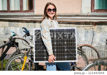 Happy woman holding solar panel, standing beside several parked bikes. Happy female wearing sunglasses and plaid shirt, enjoying sunny day in urban setting. Happy woman holding solar panel, standing beside several parked bikes. Happy female wearing sunglasses and plaid shirt, enjoying sunny day in urban setting. 121447044