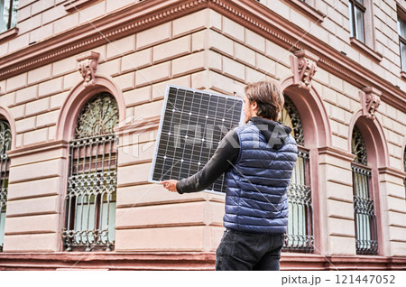 Man holding photovoltaic solar panel in front of historical building. Concept of integration of sustainable renewable energy sources into architecture. Man holding photovoltaic solar panel in front of historical building. Concept of integration of sustainable renewable energy sources into architecture. 121447052