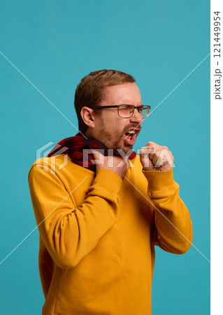 Portrait of young man in glasses suffering from sore throat and cough, warming neck with scarf against blue studio background. Portrait of young man in glasses suffering from sore throat and cough, warming neck with scarf against blue studio background. 121449954