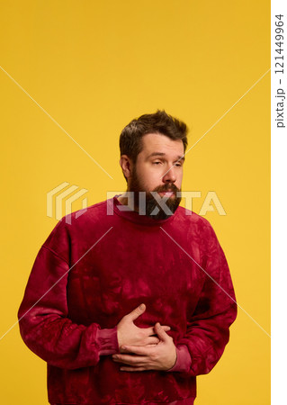 Young man feeling bad, holds hands on stomach with his hands, writhing in pain and nausea against yellow studio background. Young man feeling bad, holds hands on stomach with his hands, writhing in pain and nausea against yellow studio background. 121449964