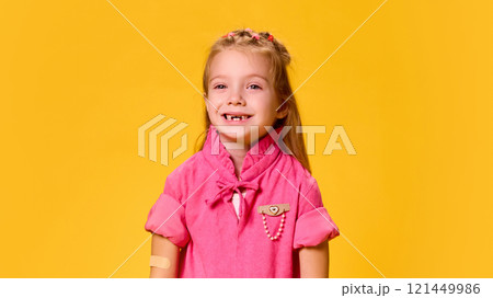 Little charming girl in pink dress smiling her arm with patch on arm after successful vaccination against yellow studio background. 121449986