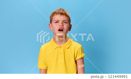 Boy in yellow shirt and with bandage on arm looking confused with furrowed brows, showcasing curiosity against blue studio background. Boy in yellow shirt and with bandage on arm looking confused with furrowed brows, showcasing curiosity against blue studio background. 121449991