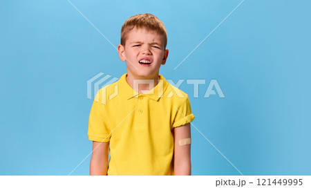 Boy grimacing slightly, bandaged arm visible, creating expressive moment of exaggerated discomfort against blue studio background. 121449995